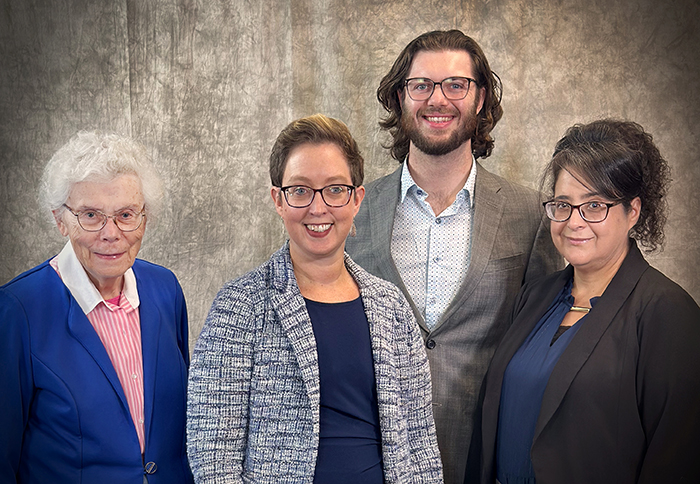 Standing in a row, an older woman with white hair wearing glasses and a blue blazer, a woman with short brown hair wearing glasses and a gray blazer, a tall man with long brown hair and a beard wearing glasses and a gray blazer, and a woman with long dark hair wearing glasses and a black blazerThree women and one man, all wearing glasses and blazers