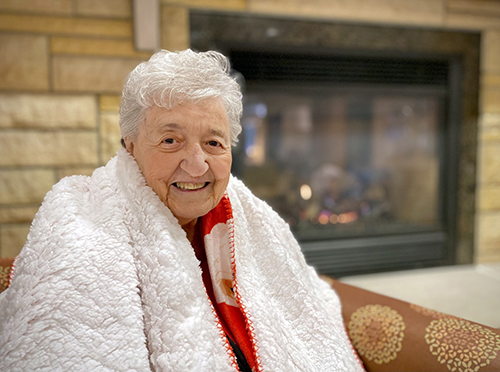 Elder woman with white hair sits with a white blanket around her shoulders in front of a fireplace.