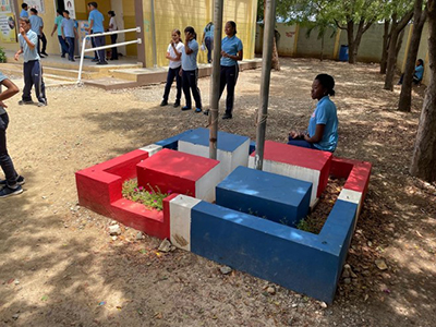 Children standing around a dirt schoolyard surrounded by a wooden fence with red and blue concrete seating cubes in the center